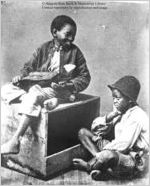 [Photograph of African American male youth sitting jauntily on a box while his companion on the floor, another African American male youth, eyes him angrily]