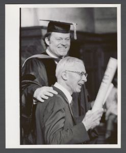 Commencement, 1970s - Smiling man holds piece of paper while professor puts an arm around him