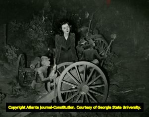 Posed photograph of woman standing over Civil War field gun, in the Civil War diorama and cyclorama painting at the Atlanta Cyclorama in Grant Park, Atlanta, Georgia, 1950s?