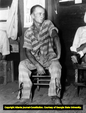 Prisoner, seated, wearing leg-irons ("picks") on each leg. Behind him is a calendar for August 1948.