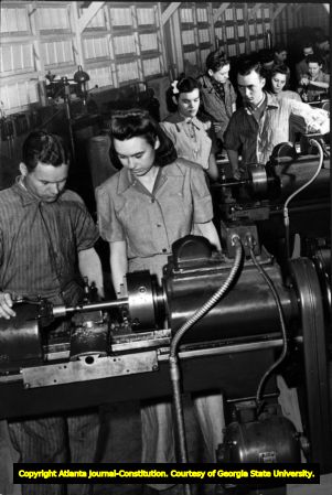 Defense industry assembly line workers. Row of metal lathe stations; at each station is a male and female worker. Photograph from 1942?
