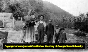 Standing on a dirt road with a mountain cabin in the background, a widow holds her infant, while five children stand at her side; her husband was killed in a shooting. Georgia mountains, ca. 1930s.