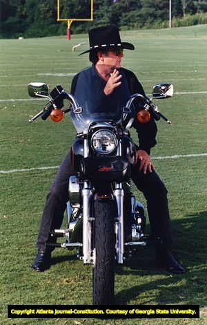 Falcons coach Jerry Glanville sits on a motorcycle on a football field for a video, 1991