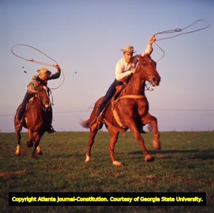 Two cowboys on horseback race across a field, 1965