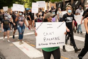 A protester marches through the street holding a sign reading 'I Can't Breathe #BlackLivesMatter #SayTheirNames #George #Taylor #Ahmaud'
