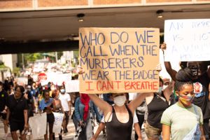 A protester holds up a sign reading 'Who Do We Call When The Murderers Wear The Badge? I Can't Breathe'