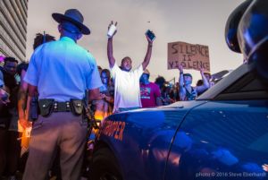 A protester standing right in front of a police car and officers holds both their hands in the air