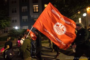 A protester marches through the streets carrying a large Industrial Workers Union Flag