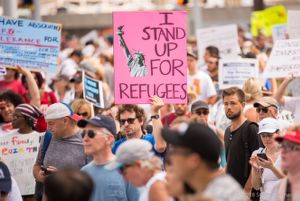 A protester holds up a pink sign reading 'I Stand Up For Refugees'