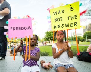 Two Children hold signs decorated with feathers and streamers reading 'Freedom' and 'War is not the Answer'.