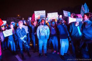 Protesters stand gathered around a protester speaking into a bullhorn