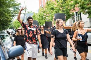 Protesters with taped mouths march by, holding hands and blacked out signs in the air