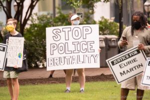 A protester wearing her mask holds a large sign reading 'Stop Police Brutality'.