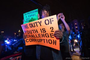 Protesters hold up signs reading 'The Duty Of Youth is 2 Challenge Corruption' and 'I'm Protesting a Rapist, Not A President'