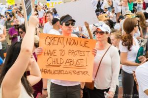 A protester holds up an orange sign reading 'Great, Now We Have to Protest Child Abuse! #FamiliesBelongTogether'