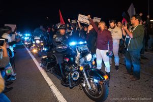 Protesters stand on either side of the street as police officers ride through the middle of them