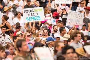 A protester among a crowd holds up a sign reading 'No One Chooses to be a Refugee'