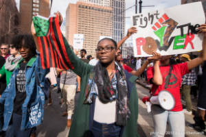 Protesters walk with American flag in black, read, and green, and sign, "Fight the Power"