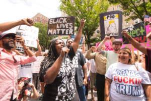 Protesters speak on a microphone and hold signs, including "Black Lives Matter" and "Poo Politician"