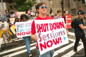 Protesters walk with signs, including "Shut down sessons now!"