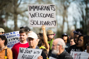 A person in an outdoor crowd holds a sign that reads, "Immigrants Welcome Solidarity"