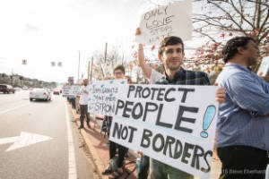 A man standing on a sidewalk holds a sign that reads, "Protect people! Not Borders"