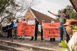 A person speaks into a microphone in front of banners at an outdoor rally