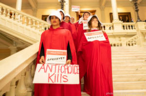 Protesters dressed as characters from Handmaiden's Tale stand on stairs in two columns holding signs, "Anti-chioce kills"