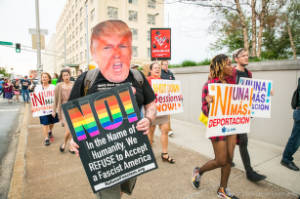 Protesters walking with signs, including Trump's face with a red X and NO written on it, "NO! In the name of Humanity, We REFUSE to Accept a Fascist America"