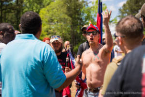 A shirtless man with a Confederate flag tattoo speaks to a group of people