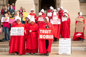 Protesters dressed as characters from Handmaiden's Tale hold signs, including "Turst Women"