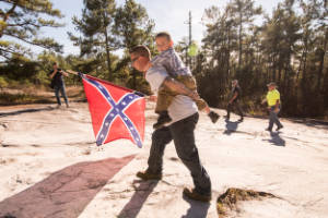A man holding a confederate flag walks up Stone Moutain with a child on his back