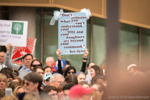 A protester holds a sign in crowd, "Don't criticize what you can't understand, your sons and your daughters are beyond your command - Bob Dylan"