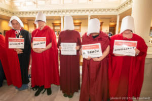 Protesters dressed as characters from Handmaiden's Tale hold signs, including "HB481 will responsible for killing women & girls"
