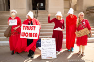 Protesters dressed as characters from Handmaiden's Tale with signs, including "Trust women" and "100% of pregnancy are caused by men. Regulate your own bodies!"