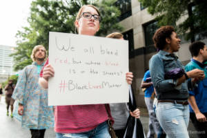 A protester holds a sign that reads, "We all bleed red, but whose blood is in the streets?"