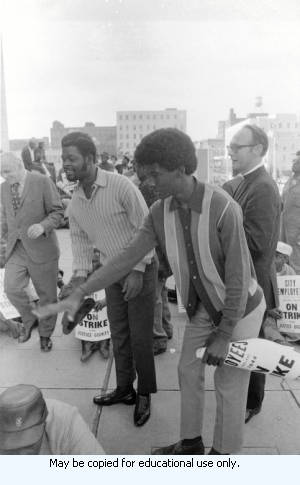 Protest organizer addressing Atlanta Garbage Strike protestors on sidewalk in front of Atlanta City Hall, downtown Atlanta, Georgia, March 31, 1970. One in a series of images labeled "Strike."