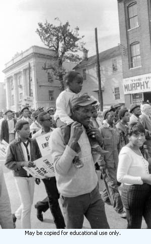 Sanitation workers strike supporters marching on Auburn Avenue, in downtown Atlanta, Georgia, March 31, 1970. Man carrying child on shoulders. Photograph is part of a series labeled "Strike."