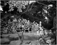 Staff eating on paper plates, woman playing an accordion, on stone steps at Davison-Paxon Company's picnic in Chastain Park (formerly, North Fulton Park), Atlanta, Georgia, July 1, 1954.