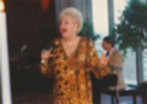 Woman singing, with man accompanying on piano in background