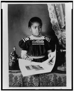 [African American girl, half-length portrait, standing at table with illustrated book, facing slightly right]
