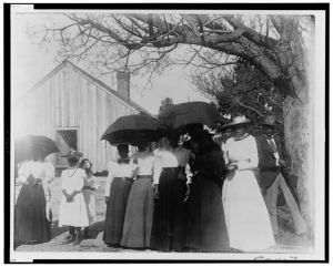 [African American women holding umbrellas to provide shade from the sun, with two men, and with a building (church or meeting house) in the background]