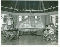 Photograph of six children in wheelchairs watching birds inside of a large cage in the recreation room of the children's pavilion at the Georgia Warm Springs Foundation, Warm Springs, Meriwether County, Georgia, 1950-1960?